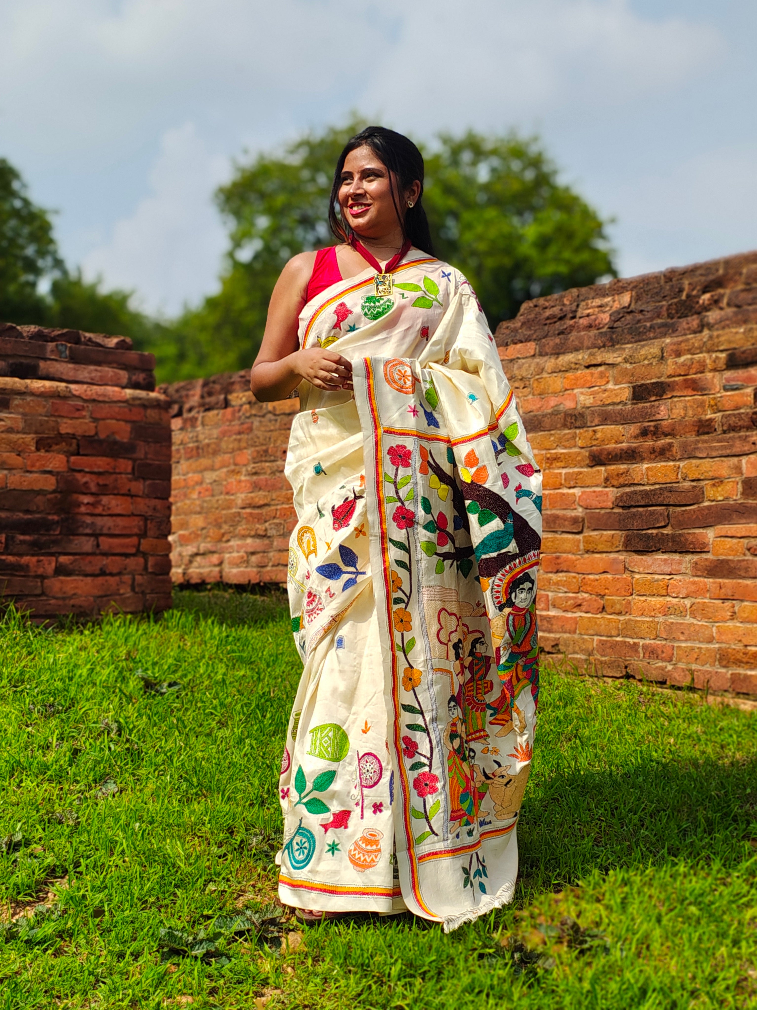 Woman in a traditional embroidered saree standing in front of ancient brick walls and greenery